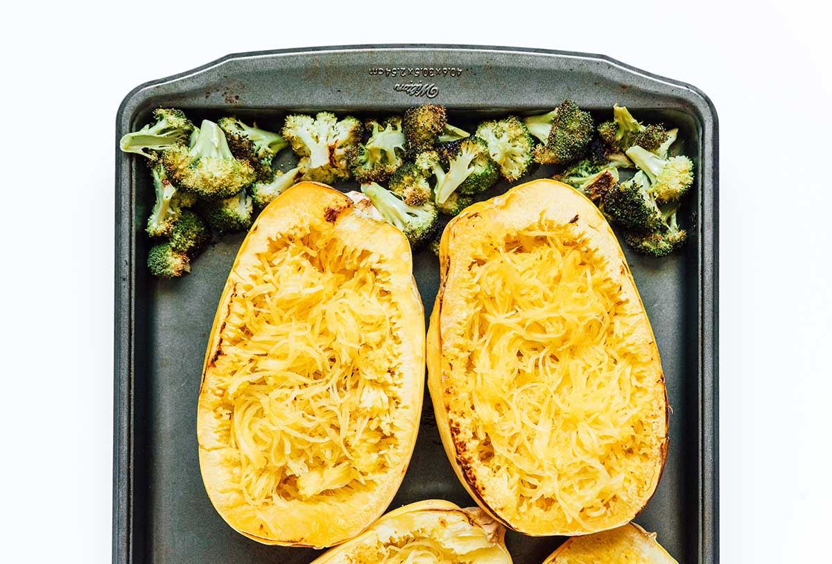 Two cups of broccoli and two spaghetti squash halves filled with squash strands on a baking sheet