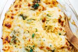 A baked spaghetti squash alfredo casserole dish with melted cheese, broccoli, and a wooden spoon scooping out a portion.