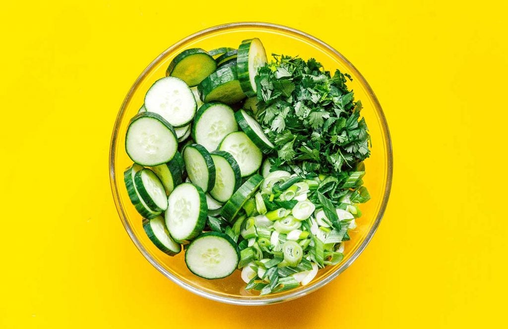 A glass bowl filled with cucumber slices, chopped green onion, and chopped cilantro