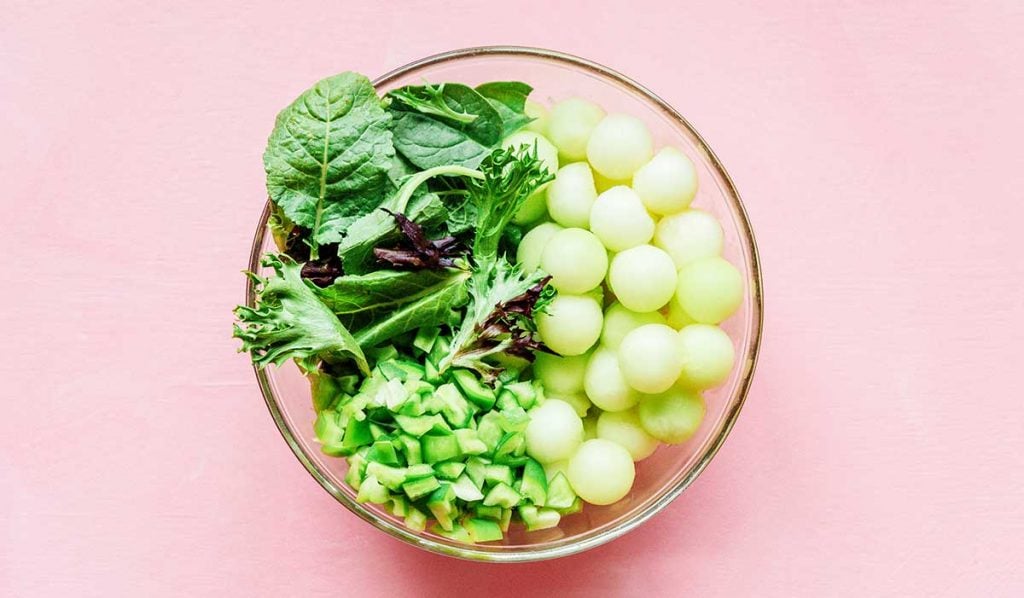 A glass bowl filled with spring mix, bell pepper, and honeydew melon balls