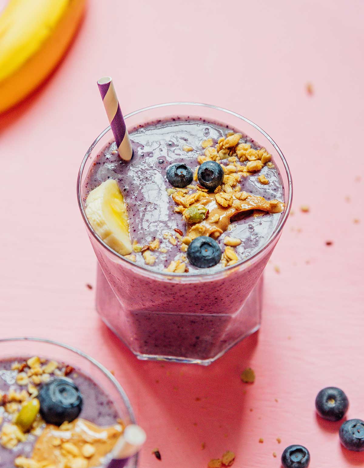 A glass filled with blueberry banana smoothie and topped with nut butter, granola, blueberries, and a banana slice