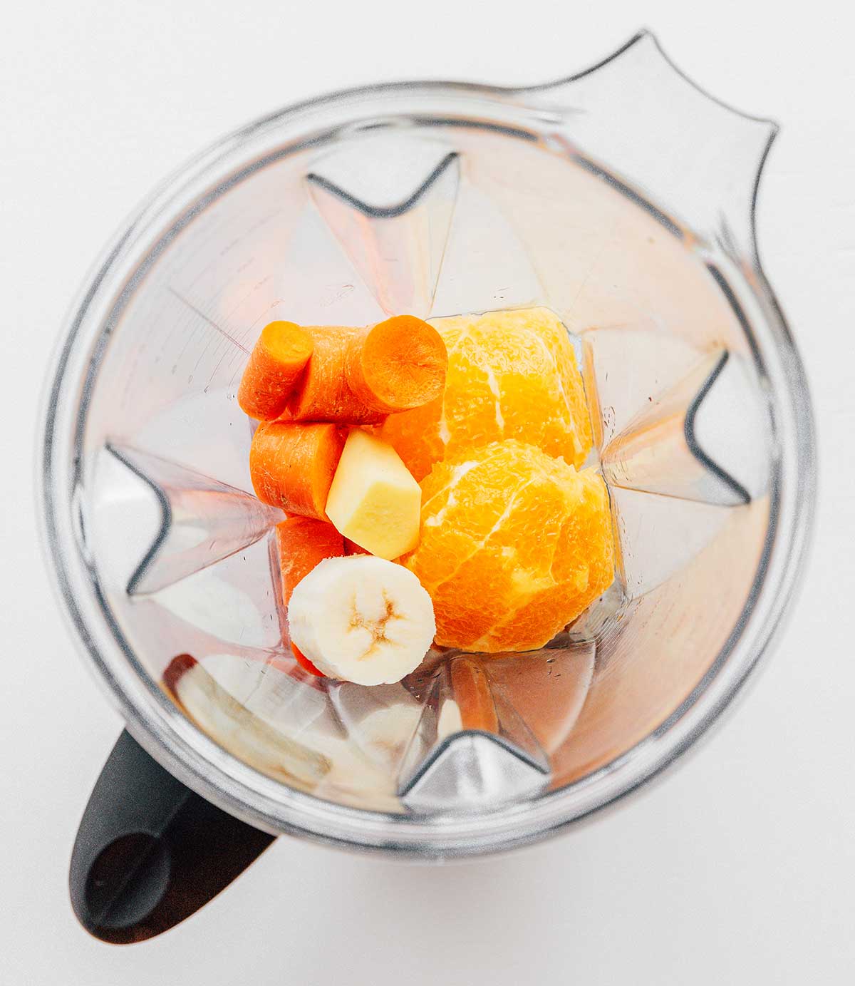A blender filled with sliced banana, sliced carrots, chopped pineapple, and 2 halves of an orange