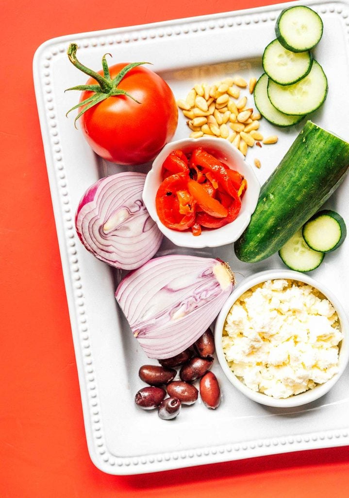 A white platter filled with a tomato, pine nuts, English cucumber slices, a halved red onion, feta cheese, and Kalamata olives