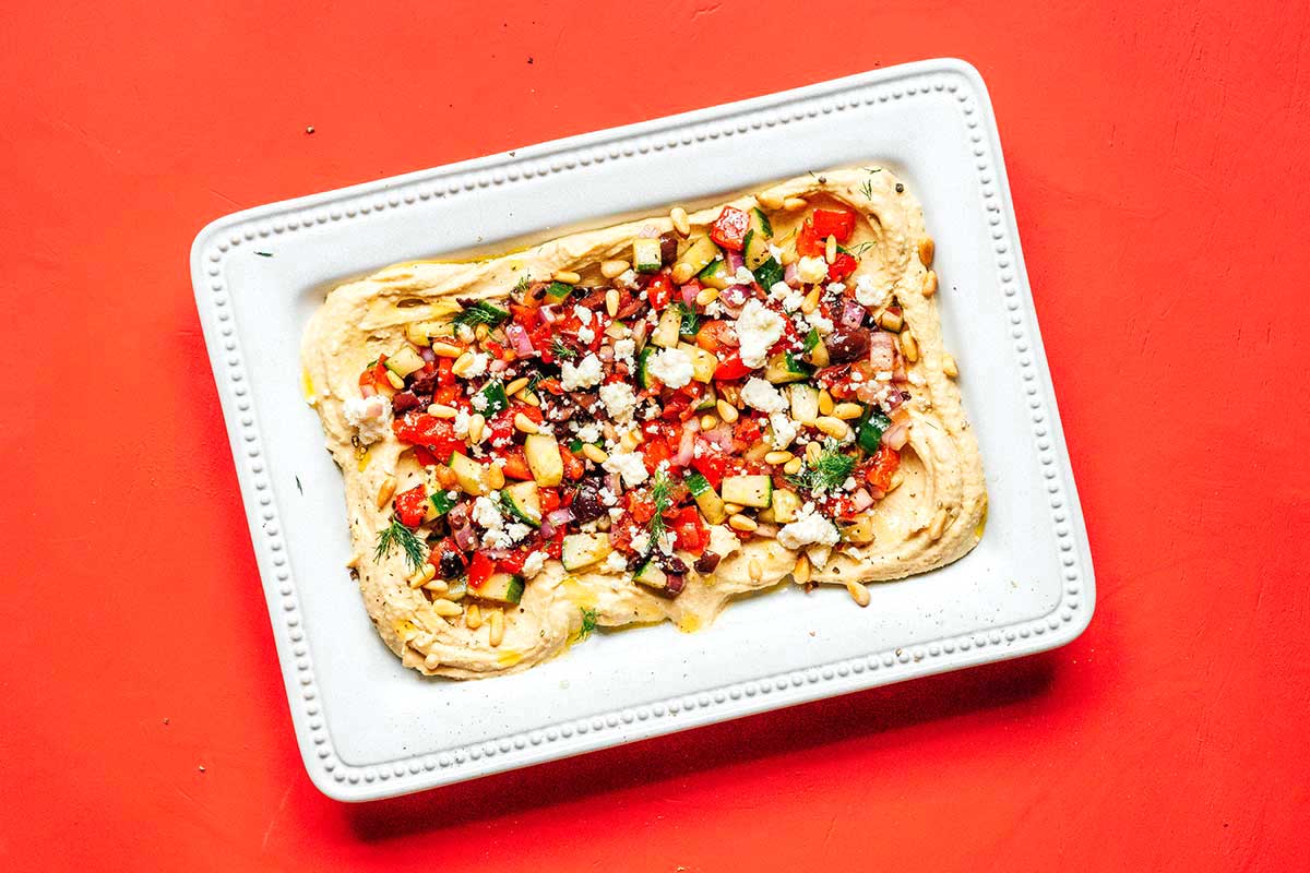 A white rectangular tray coated with a layer of hummus and topped with Mediterranean toppings, pine nuts, and feta cheese