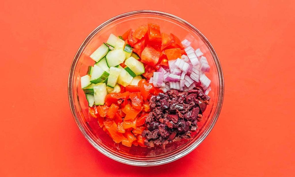 A glass bowl filled with diced cucumber, tomatoes, red onion, Kalamata olives, and roasted peppers