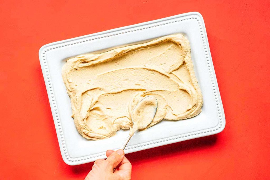 A hand using a spoon to spread a layer of hummus onto a white rectangular tray