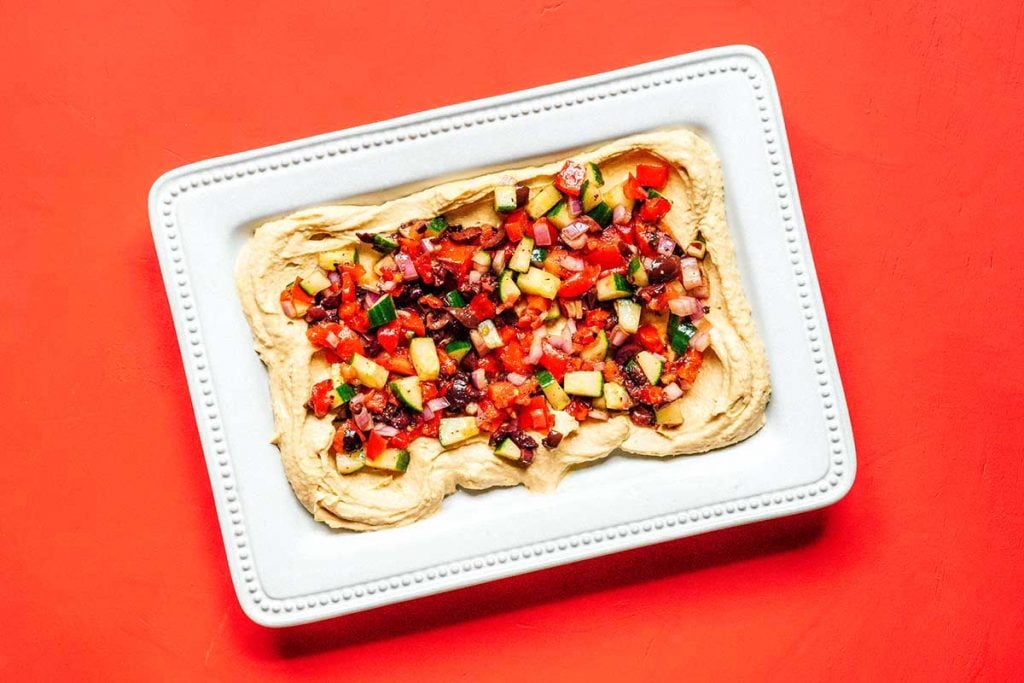 A white rectangular tray coated with a layer of hummus and topped with Mediterranean toppings