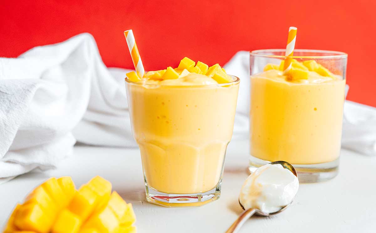 Two glasses of mango smoothie, a spoon filled with Greek yogurt, and a diced mango