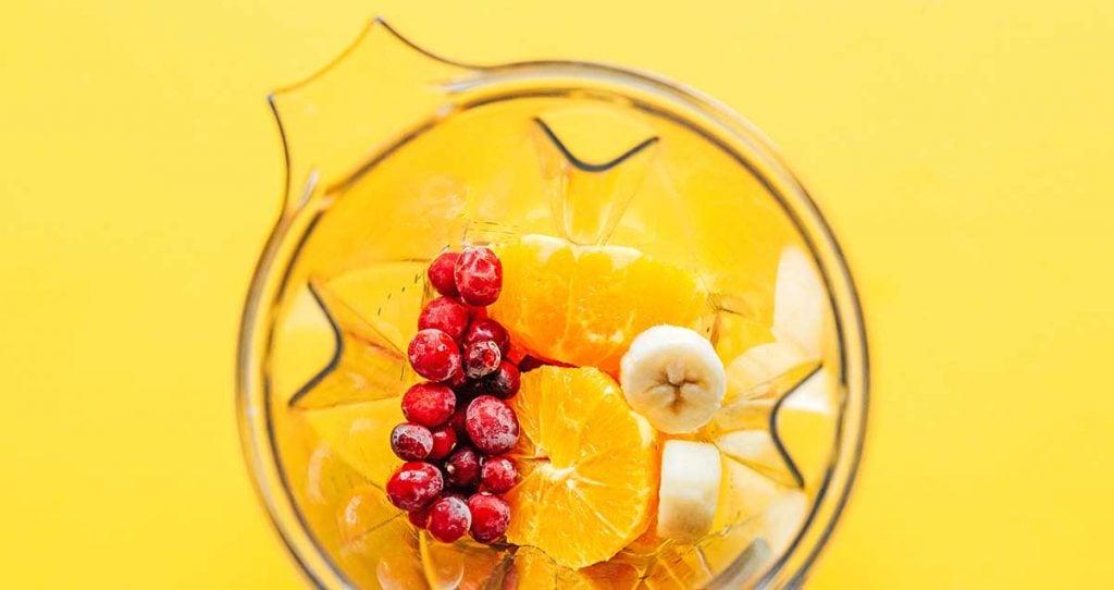 A blender filled with frozen cranberries, peeled and pithed oranges, banana slices, and orange juice
