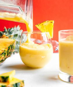 A pineapple smoothie cascades from the blender into a glass, elegantly garnished with a fresh pineapple slice. Another glass and chunks of juicy pineapple rest on a white surface, set against the vibrant orange background.