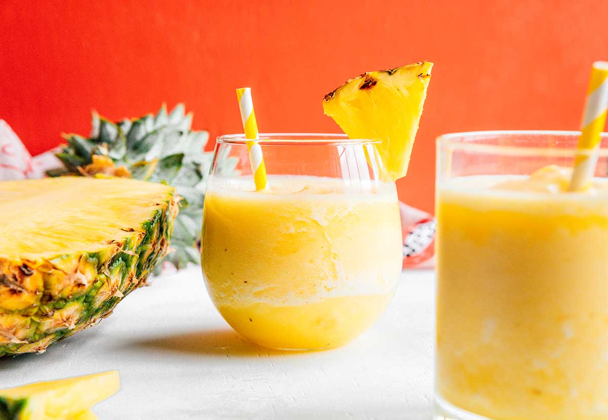 Two glasses filled with pineapple smoothie and half of a pineapple laying skin-down on a white background