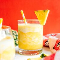 A glass of yellow frozen pineapple smoothie with a yellow-and-white straw and a pineapple wedge garnish, set against a red background. A similar drink and a fresh pineapple are visible in the background.