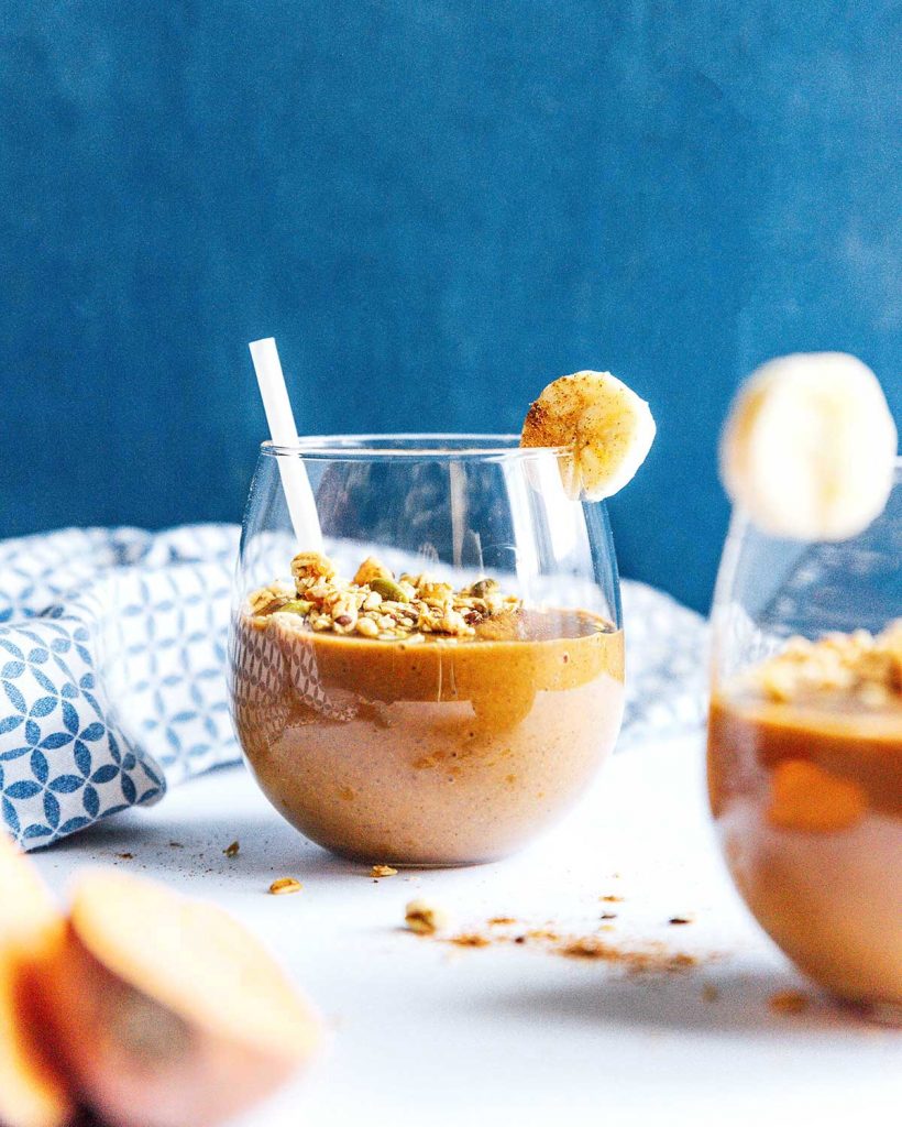 A glass filled with sweet potato smoothie and topped with seeds, granola, ginger, and a slice of banana
