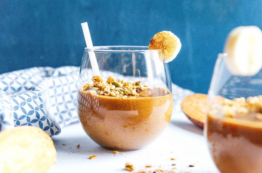 A glass filled with sweet potato smoothie and topped with seeds, granola, ginger, and a slice of banana