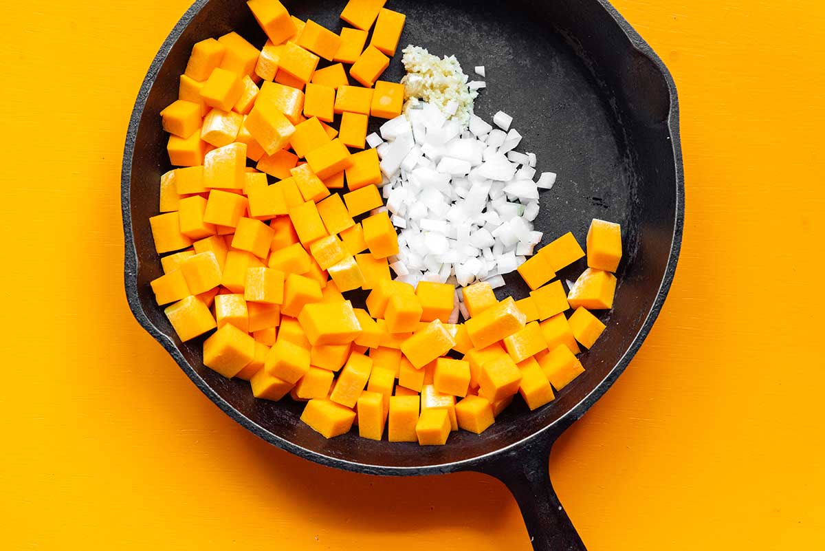 A cast iron skillet filled with diced butternut squash, chopped onion, and minced garlic