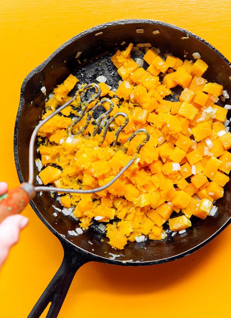 A potato masher mashing the diced butternut squash in a cast iron skillet