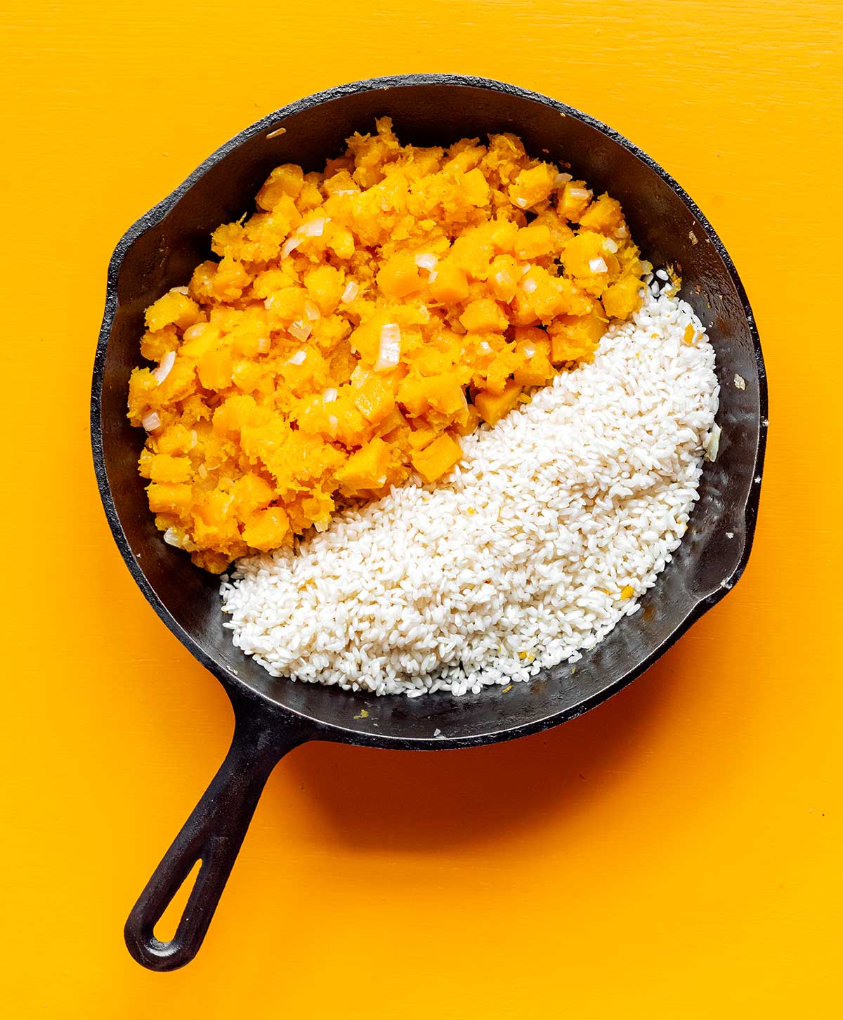 A cast iron skillet filled with mashed butternut squash, chopped onion, and Arborio rice