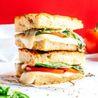 Stacked caprese panini with melted cheese, tomato slices, and basil between thick bread slices against a bright red background, garnished with loose basil leaves.