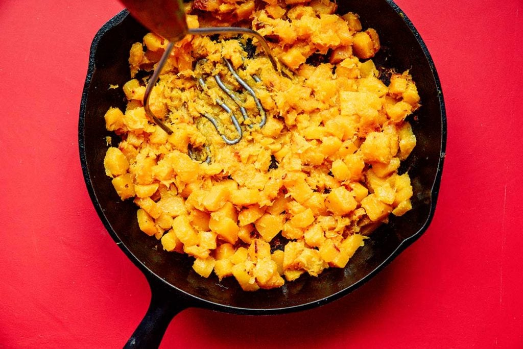Mashing cooked butternut squash with a potato masher in a cast iron skillet