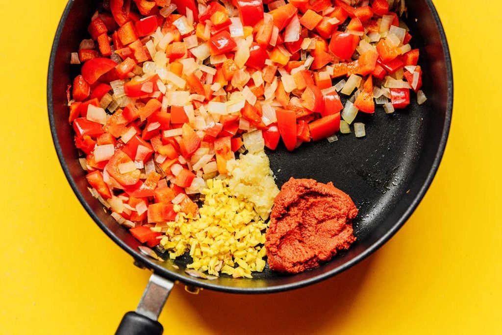 A blend of diced white onion and red bell pepper in a sauté pan alongside red curry paste, ginger, and garlic