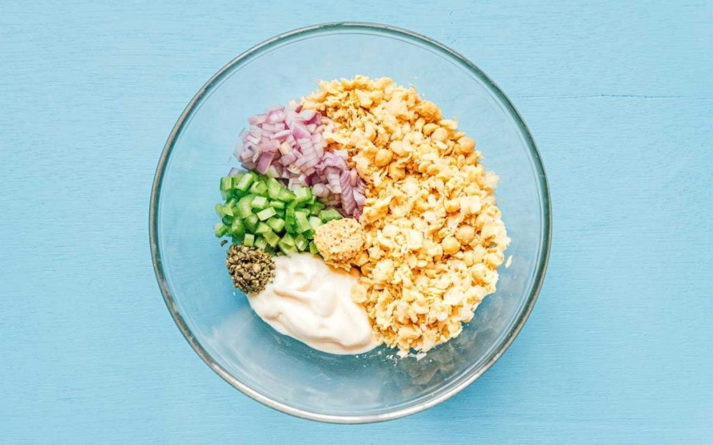 A bowl filled with diced red onion, celery, and capers, mashed chickpeas, mayonnaise, and dijon mustard