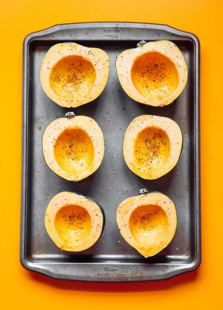 Six acorn squash halves coated in olive oil, salt, and pepper lying face up on a baking sheet 