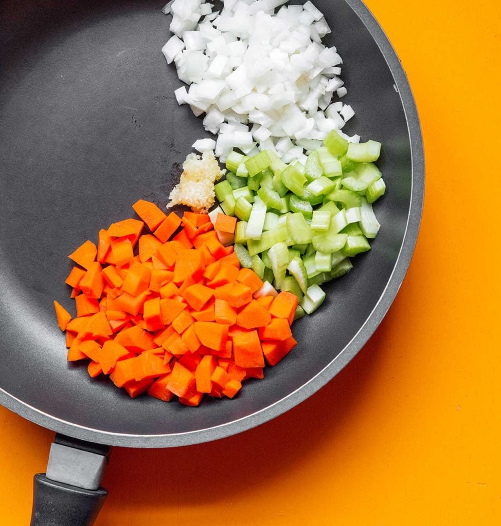 A skillet filled with chopped white onion, celery, carrots, and minced garlic