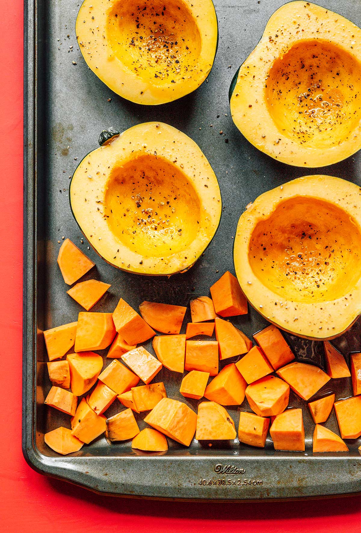 Four acorn squash halves and cubed sweet potatoes laid out on a baking sheet and topped with olive oil, salt, and pepper