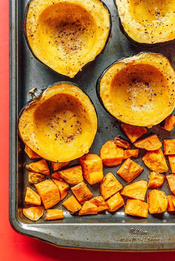 Four cooked acorn squash halves and cubed sweet potatoes laid out on a baking sheet and topped with olive oil, salt, and pepper