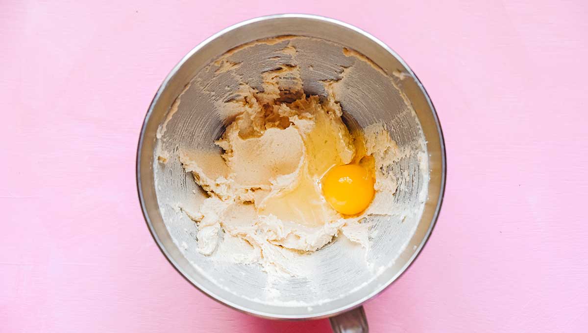 A metal mixing bowl filled with a butter and sugar mixture with an unmixed egg and vanilla on top