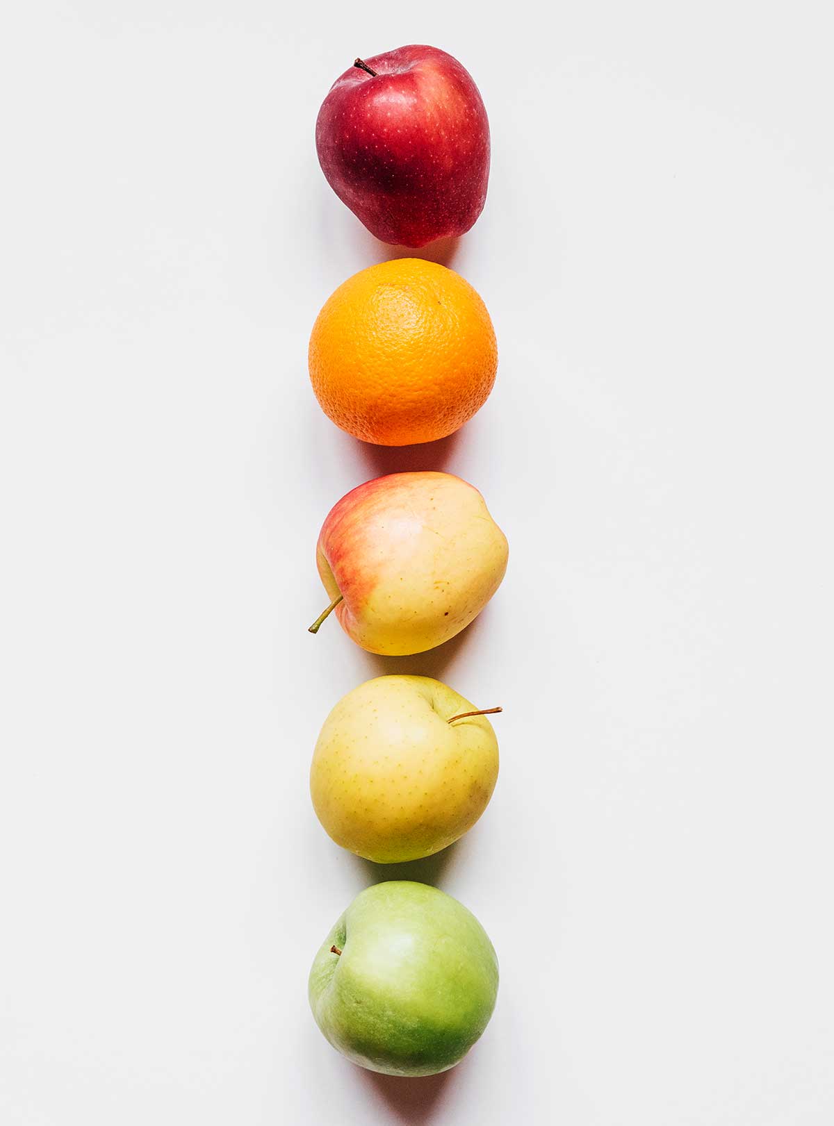 A rainbow of apples and an orange laid out in a vertical line from red to green