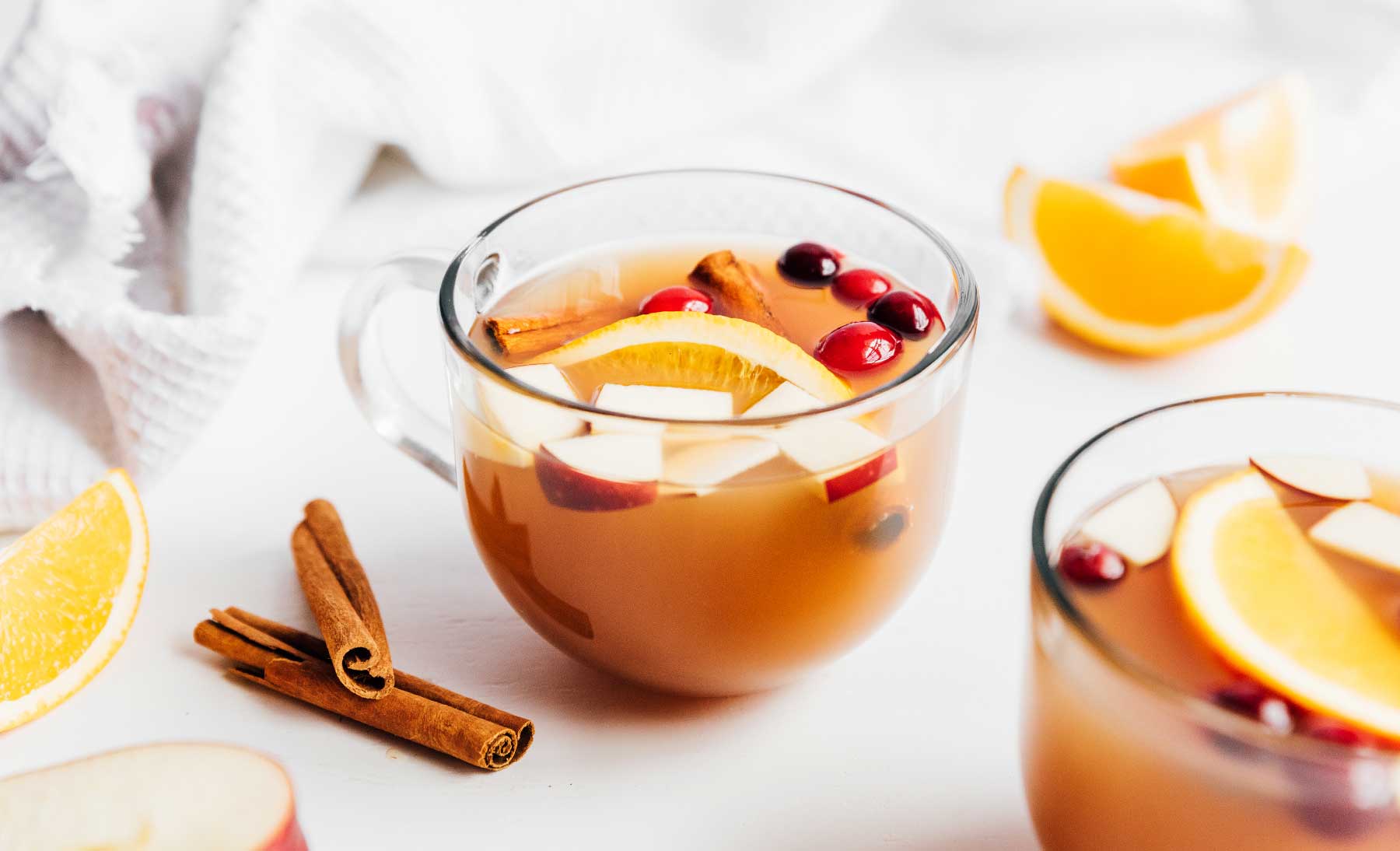 A glass of homemade apple cider garnished with cinnamon sticks, cranberries, and chopped fruit