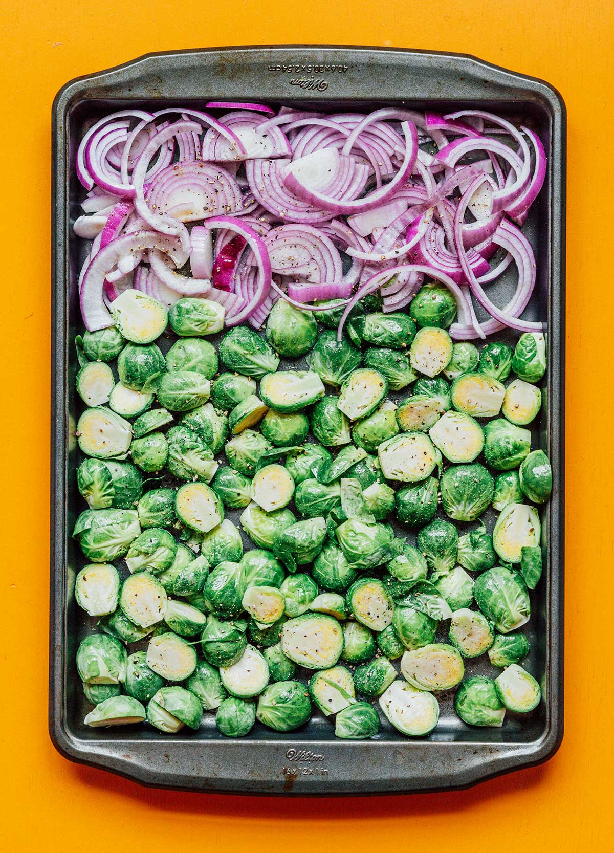 Sliced red onion and halved Brussels sprouts laid out on a baking sheet and topped with olive oil, salt, and pepper
