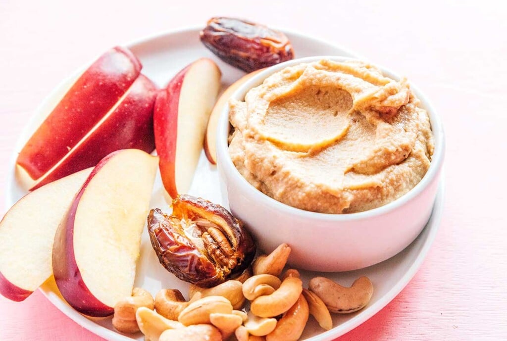 A white bowl filled with sweet cashew cream on top of a white plate surrounded by apple slices, dates, and cashews