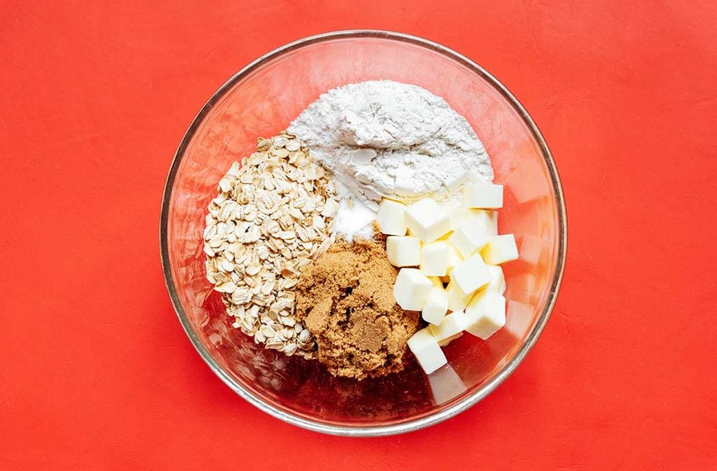 A clear glass bowl filled with all of the crisp ingredients: flour, rolled oats, brown sugar, cinnamon, salt, and unsalted plant-based butter