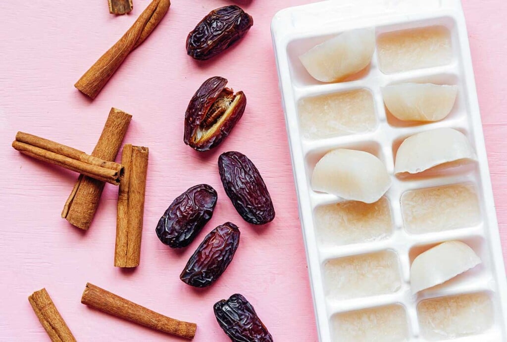 Cinnamon sticks, dates, and an ice cube tray filled with almond milk ice cubes arranged on a pink background