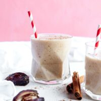Two glasses of a healthy date shake with red-striped straws, surrounded by dates and cinnamon sticks, against a pink background.