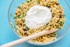 A glass bowl filled with a delightful freekeh salad, mixed with herbs and raisins, topped with a dollop of yogurt. A wooden spoon rests inside the bowl.