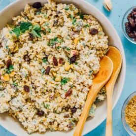 A bowl of freekeh salad with raisins, pistachios, and herbs is paired with two wooden spoons. Nearby, small bowls hold extra ingredients and lemon wedges on a blue surface.