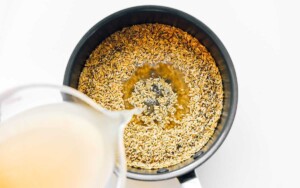 Pouring liquid from a measuring cup into a pot filled with oats on a white background demonstrates the basics of cooking. Just as one learns how to cook freekeh, mastering these steps is essential for culinary success.