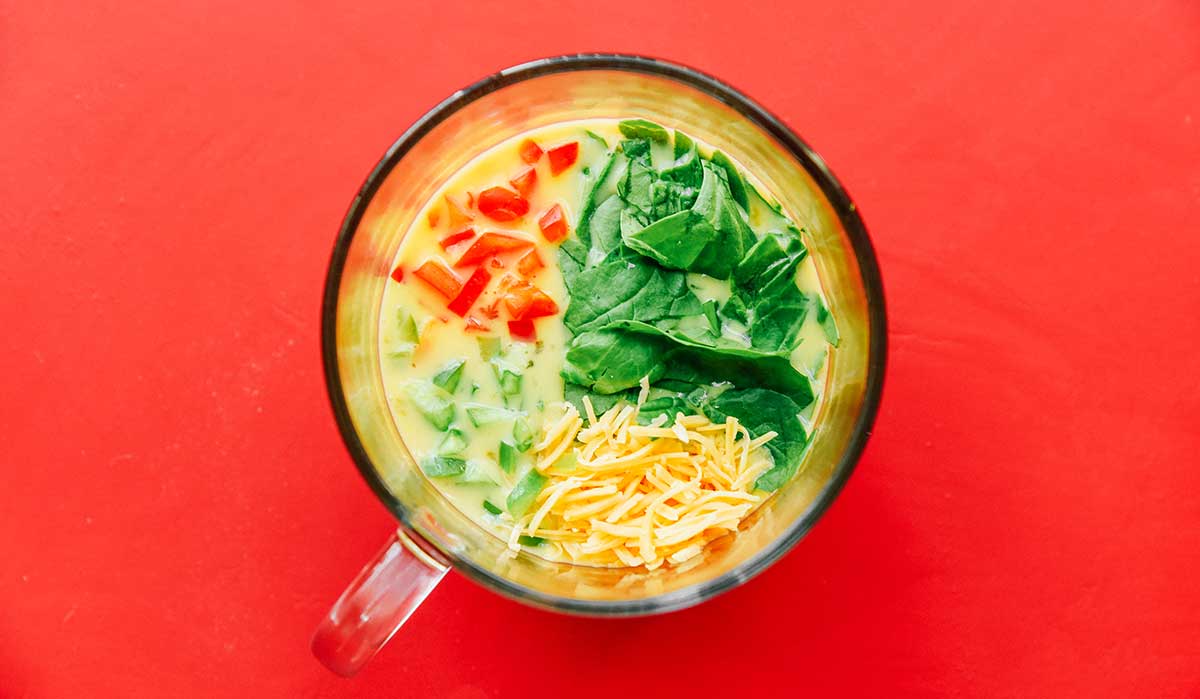 Whisked eggs and milk in a clear glass mug topped with diced red pepper, diced green pepper, chopped spinach, and shredded cheddar cheese
