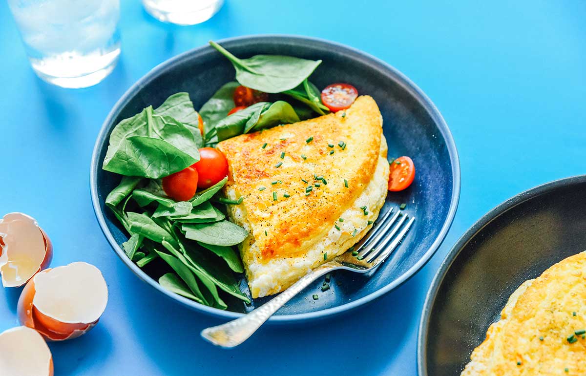 Half of a folded fluffy soufflé omelette on a plate alongside spinach and cherry tomatoes