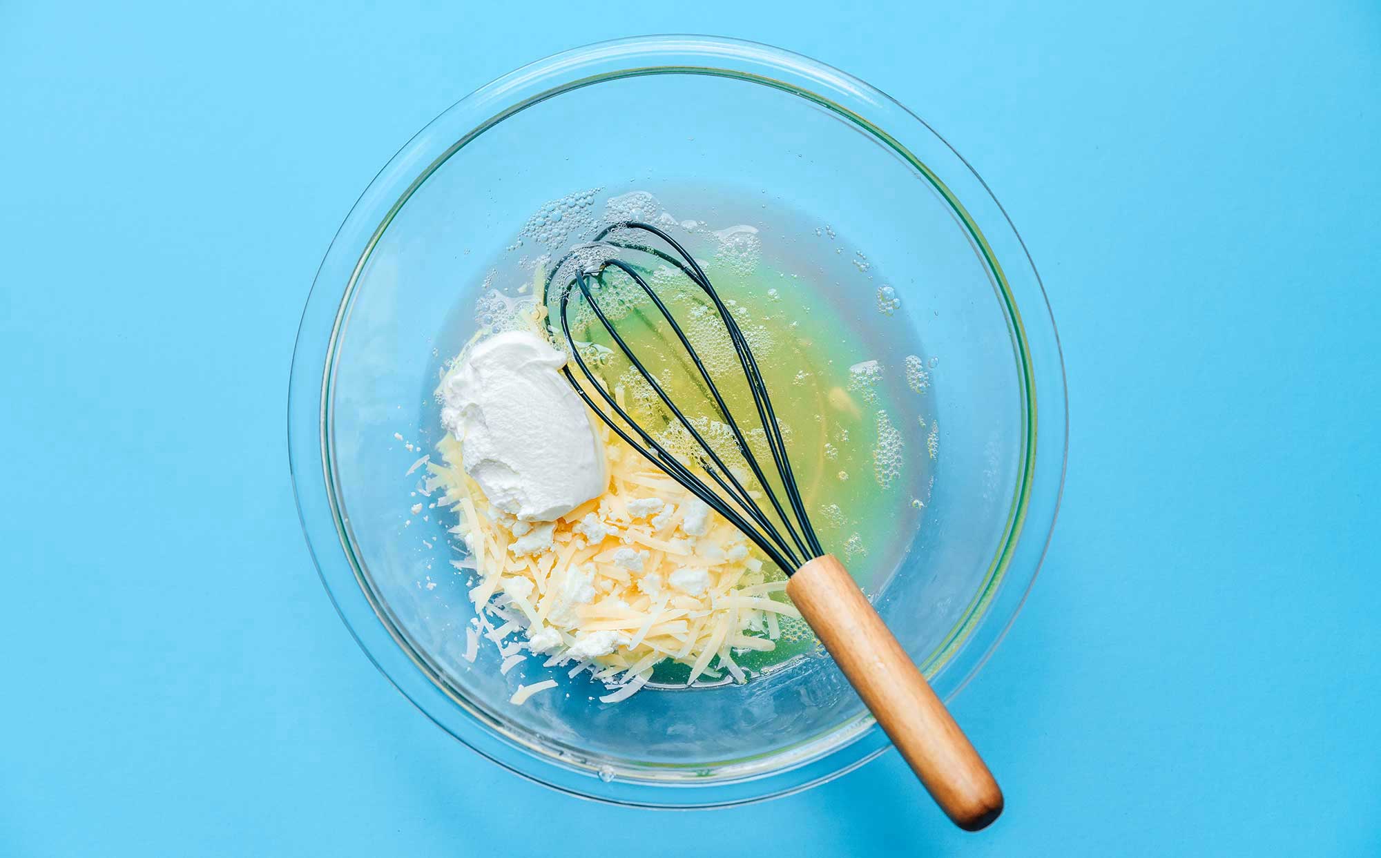 A whisk in a clear glass bowl filled with egg whites, sour cream, feta, and parmesan