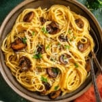 A bowl of vegetarian carbonara spaghetti topped with sautéed mushrooms, grated cheese, chopped parsley, and black pepper, with a fork resting on the side.
