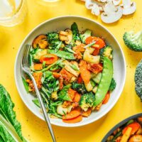 A bowl of Buddhas delight stir-fry with colorful bell peppers, snow peas, broccoli, and mushrooms on a yellow background, accompanied by sliced mushrooms and glasses of ice water.
