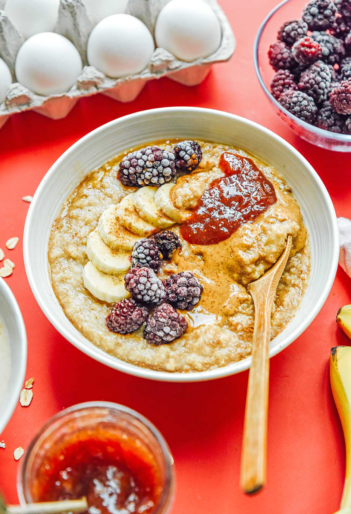 A white bowl filled with egg white oatmeal and topped with bananas, raspberries, jam, peanut butter, and brown sugar