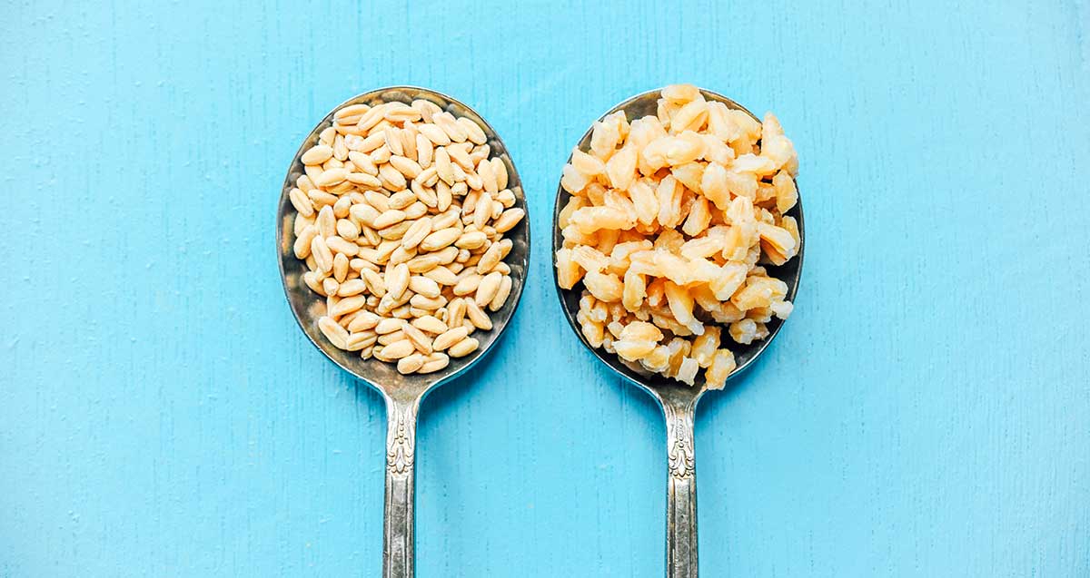 Two round metal spoons placed side-by-side, the left spoon filled with uncooked farro and the right spoon filled with cooked farro 