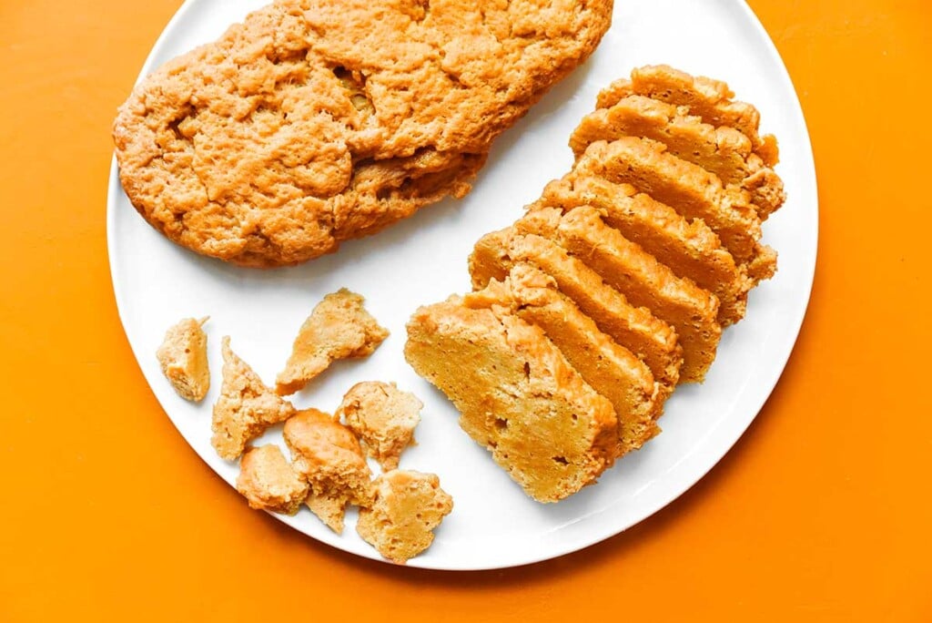 A whole and a sliced loaf of seitan sitting side by side on a white plate, along with a few pieces of cubed seitan