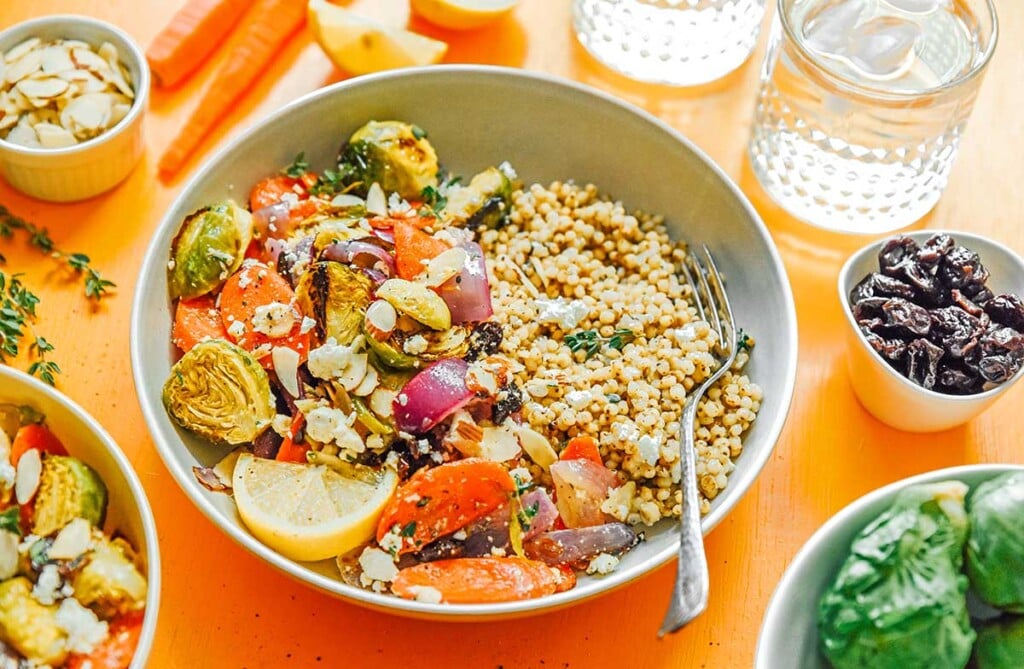 A roasted vegetable grain bowl filled with seitan, red onion, Brussels sprouts, carrots, lemon slices, feta cheese, and more