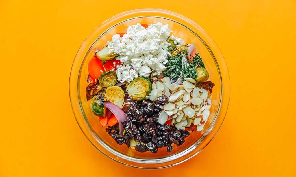 A clear glass bowl filled with roasted vegetable grain bowl ingredients like roasted veggies, feta cheese, sliced almonds, and dried cherries